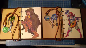 Two journals side-by-side, opened to the character artwork and showing off their personalized place markers and string closures.
