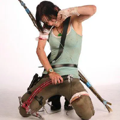A woman cosplaying as the rebooted Lara Croft, kneeling whilst adminstering first aid upon herself.